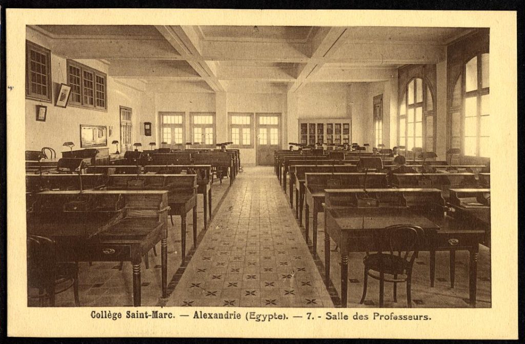"Collège Saint-Marc - Alexandrie (Egypte). -7. Salle des Professeurs" The staff room in the Collège Saint Marc contains many individual wooden desks with lamps and chairs. It is a spacious room with high ceilings.